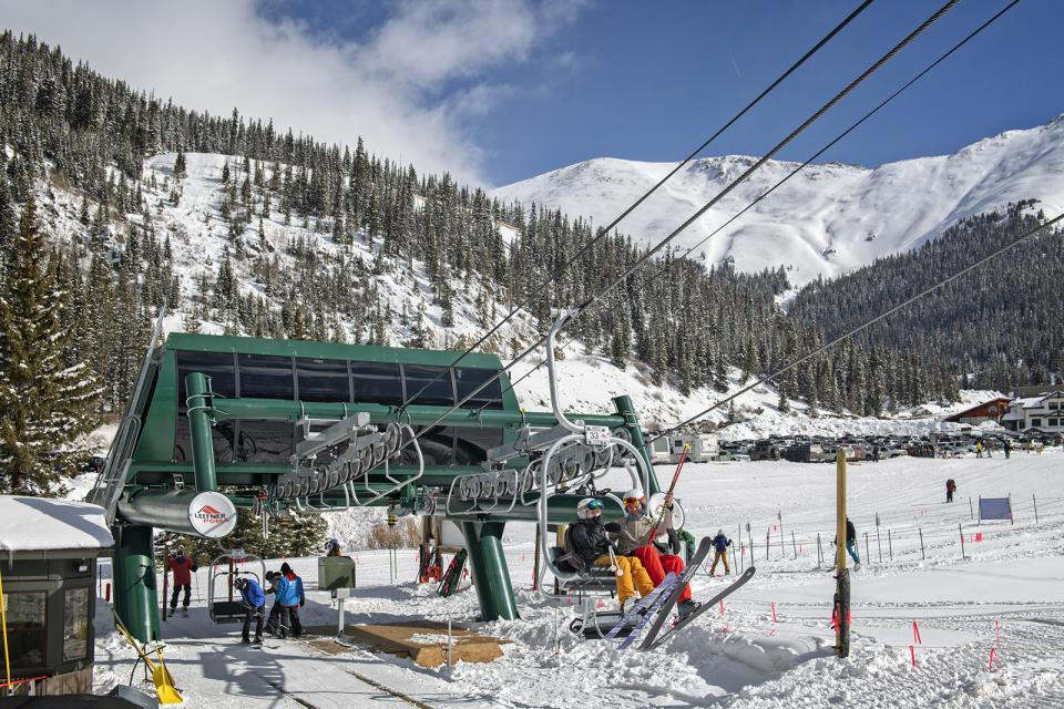 Skiers riding a Leitner-Poma chairlift departing the base terminal at Arapahoe Basin, with snow-covered mountains, pine trees, and the ski area parking lot in the background.