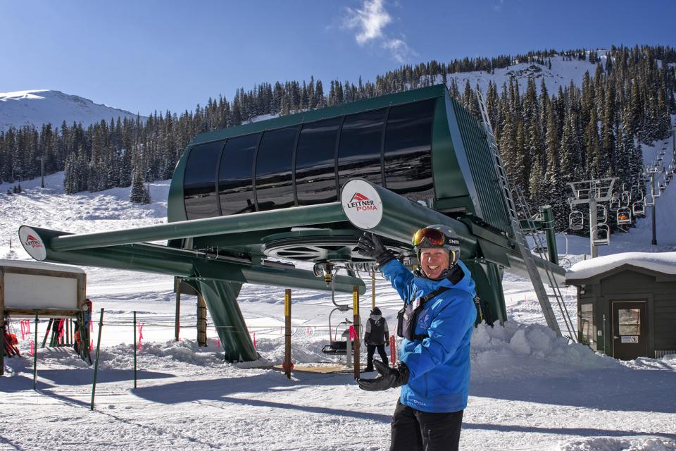 Resort operator standing in front of a Leitner-Poma chairlift terminal at Arapahoe Basin, with skiers, lift infrastructure, and snowy mountain terrain in the background.