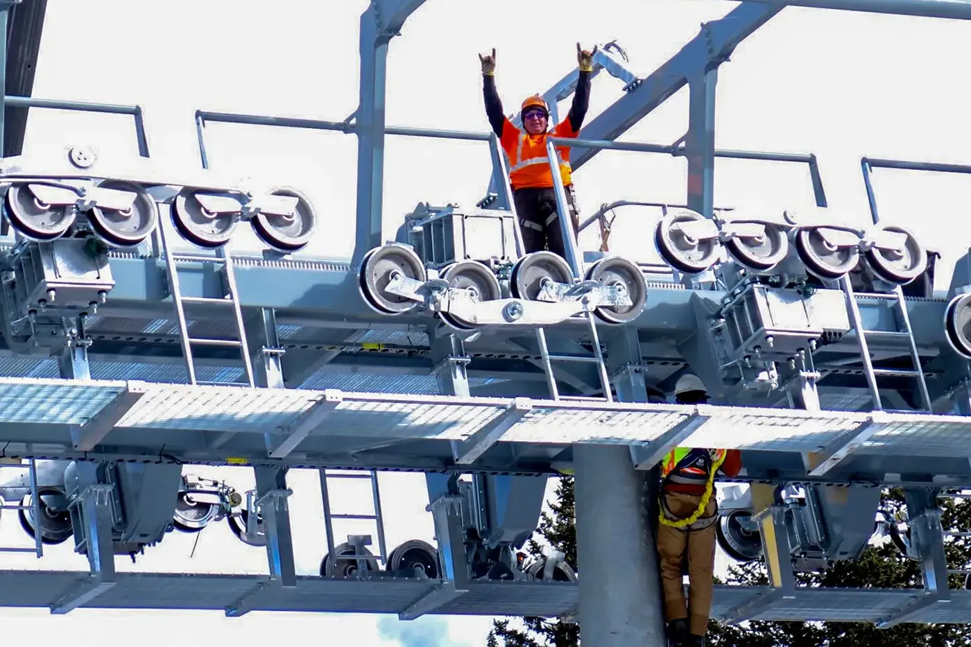 Two Leitner-Poma field technicians in safety gear work on assembling a ski lift tower. One worker stands atop the structure raising their arms in celebration, surrounded by pulleys and metal components under bright daylight.