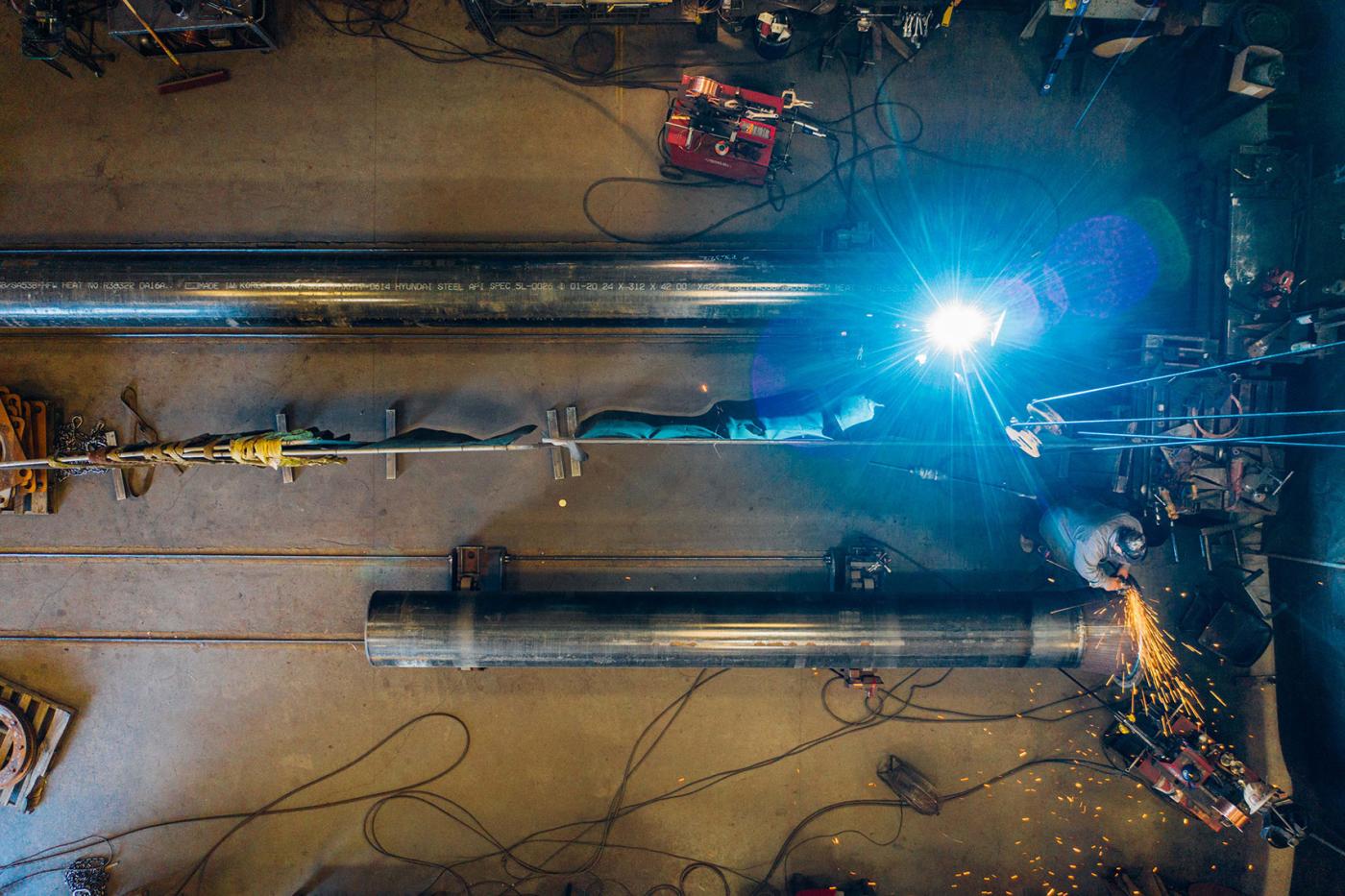 Overhead view of two Leitner-Poma workers welding and grinding large steel lift components inside a workshop, with bright sparks and light from the welding torch illuminating the workspace.